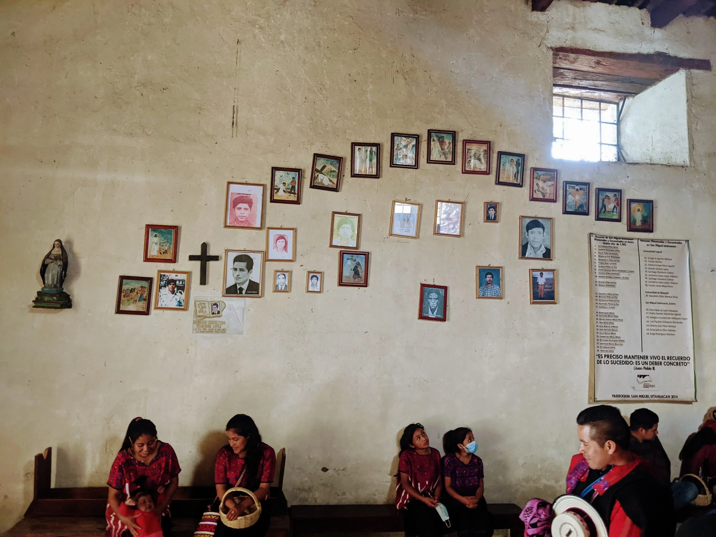 People sitting on wooden benches inside a church with white walls, several framed photos and artwork, a statue of a saint, and a window letting in natural light.