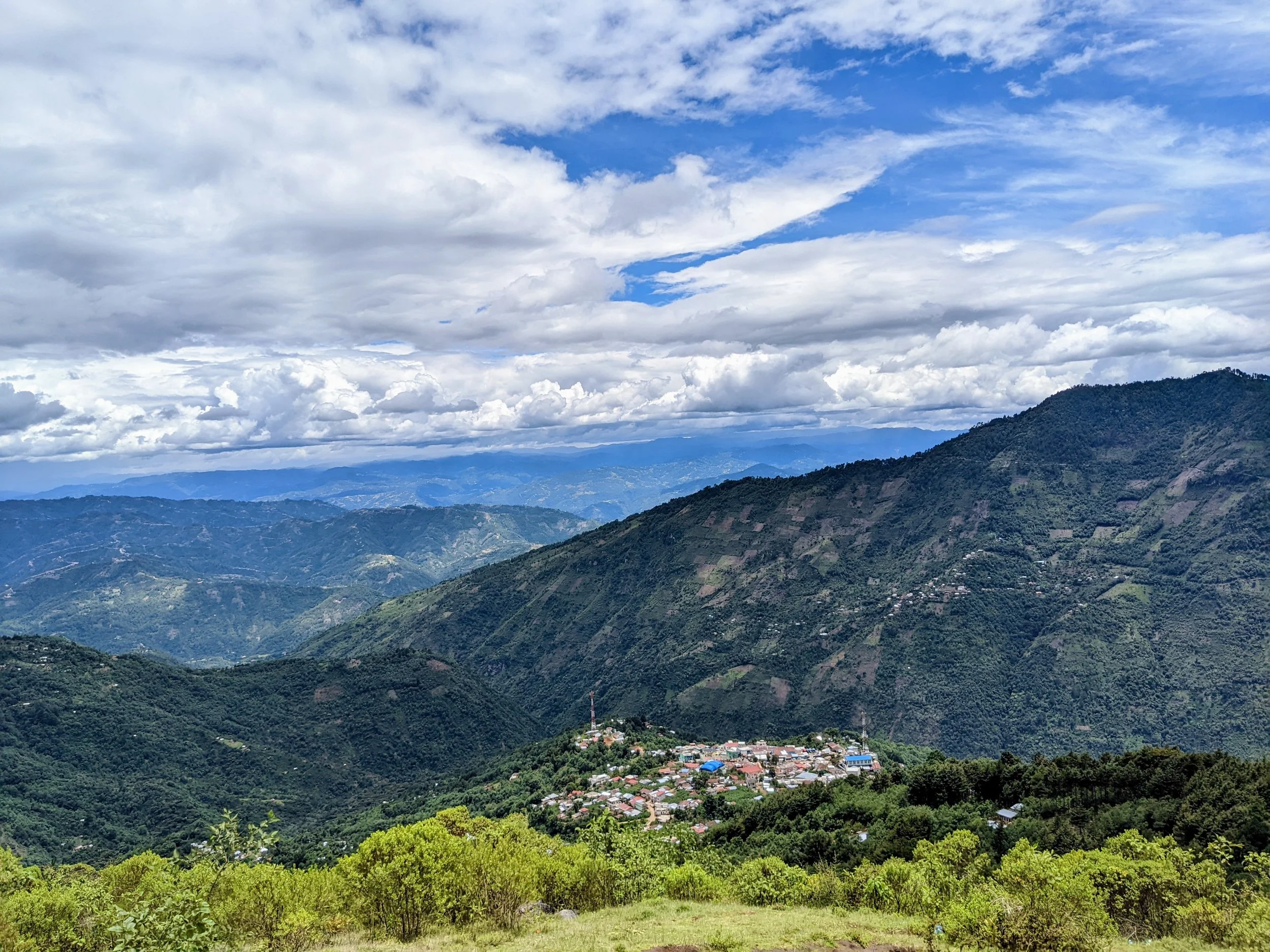 Scenic view of mountains and a small village with houses, surrounded by lush green trees under a partly cloudy sky.