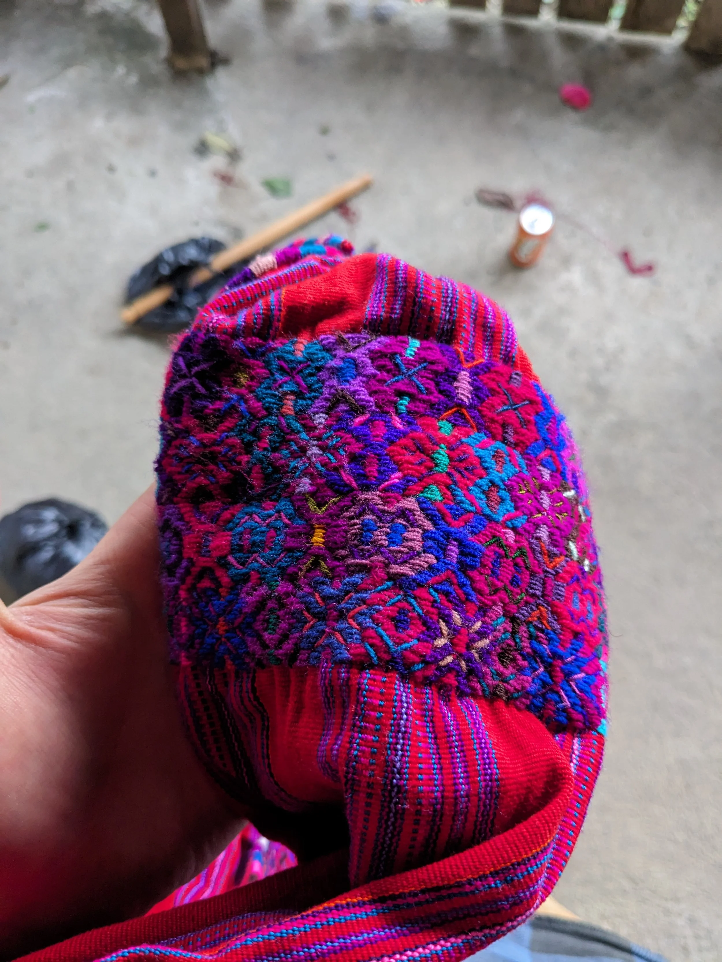 A colorful embroidered textile fabric, possibly clothing, being held by a person's hand, with a concrete floor and some scattered small objects in the background.