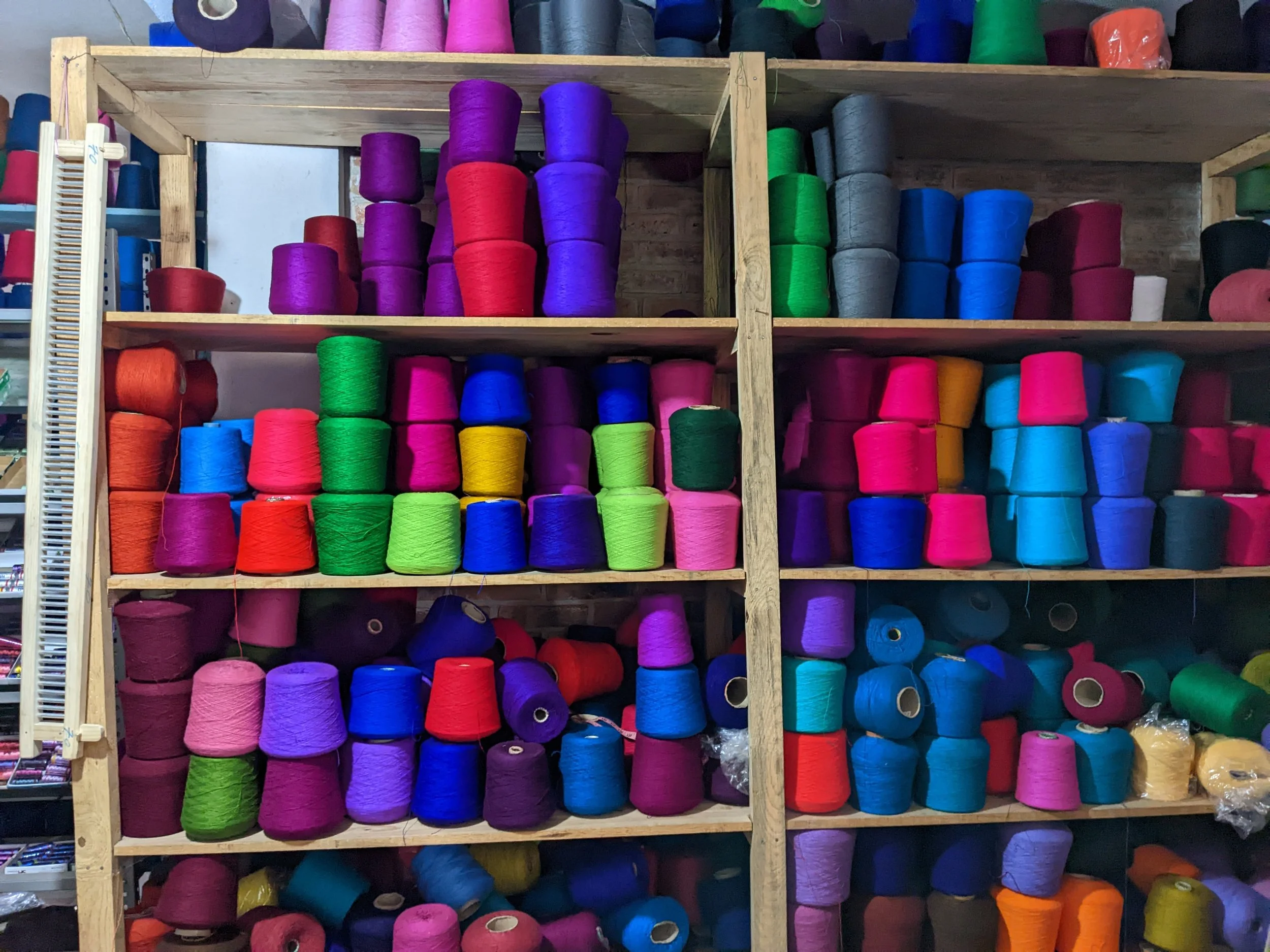 A wooden shelving unit holding large spools of colorful thread, with colors including purple, red, green, blue, pink, yellow, and gray, arranged on multiple shelves.