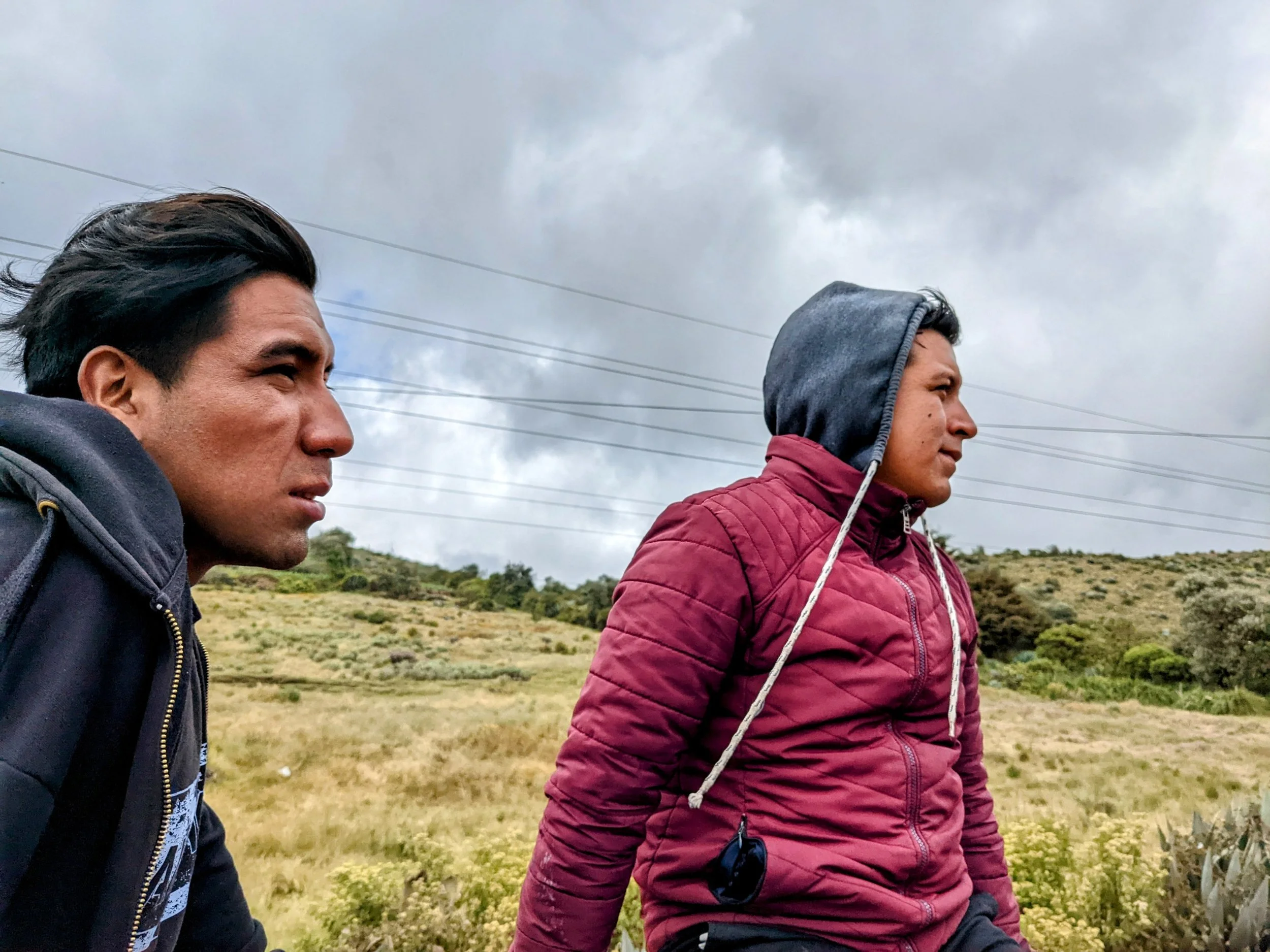 Two men are walking outdoors in a grassy and hilly landscape with cloudy skies and power lines overhead. One man has black hair and is wearing a black jacket, while the other has a gray hoodie and a maroon jacket.