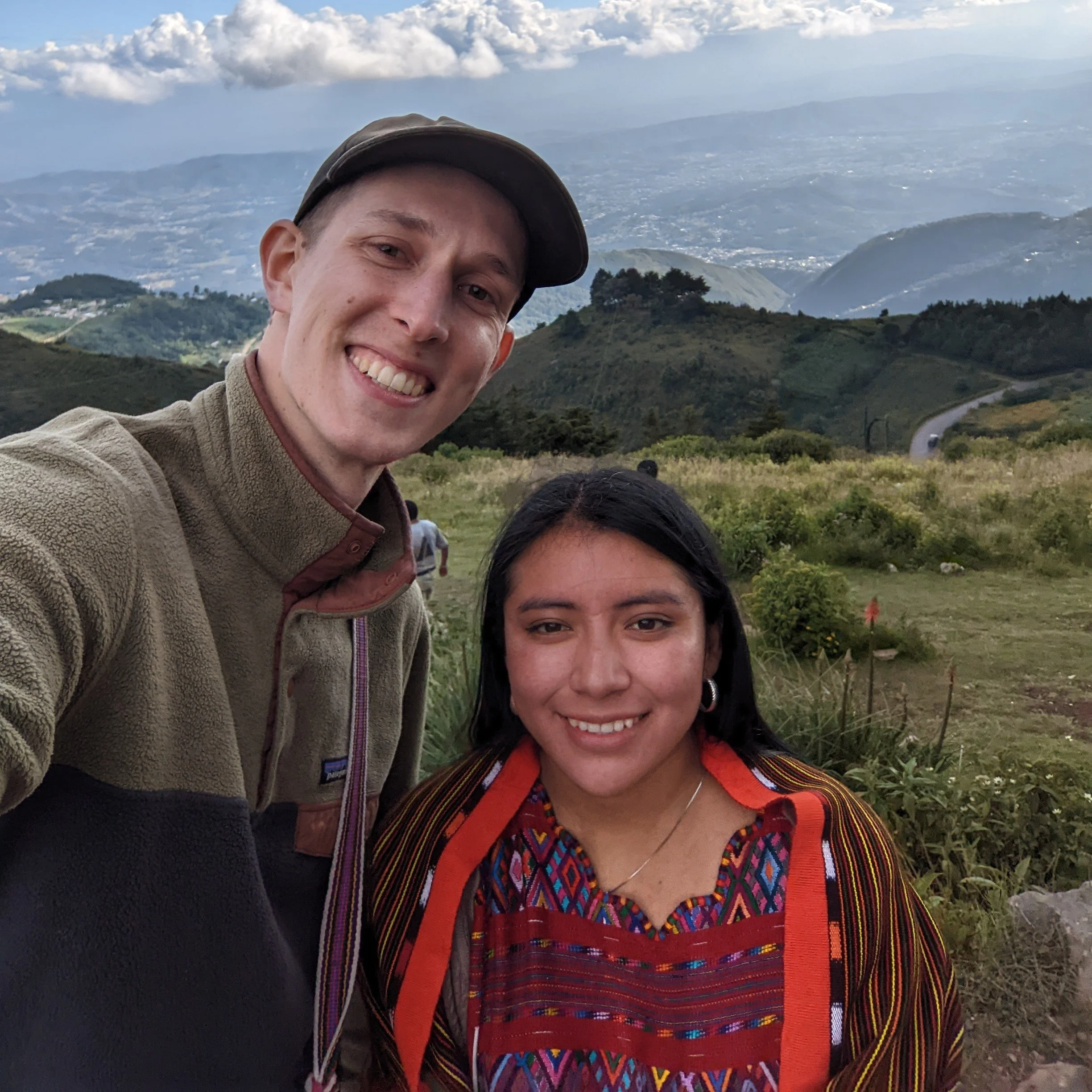 A smiling man in outdoor clothing and a hat, taking a selfie with a scenic landscape of rolling hills, mountains, and a cloudy sky in the background. A woman with dark hair and colorful traditional clothing is partially visible in the foreground.