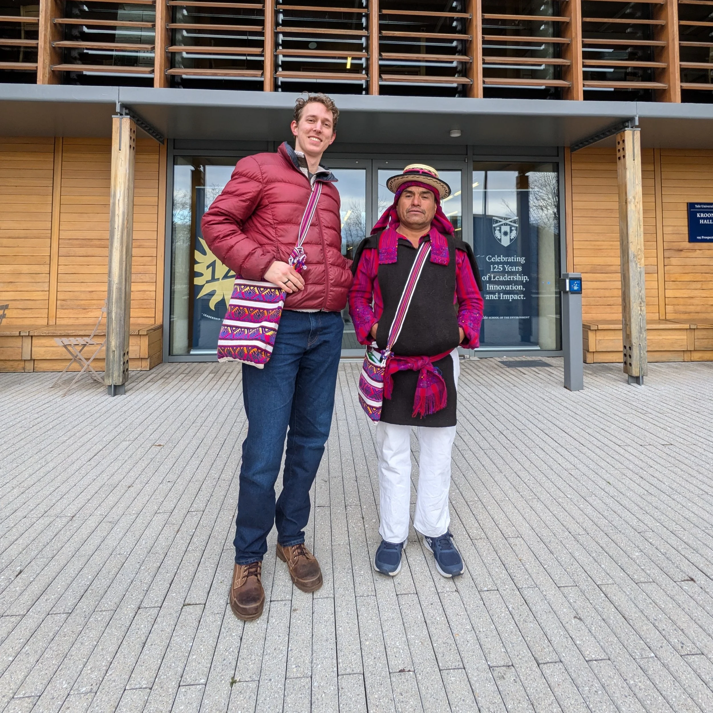 Two men standing outside in front of a modern building with wooden paneling and glass windows. The man on the left has light skin, curly hair, and is smiling, wearing a red quilted jacket, blue jeans, and brown boots. The man on the right has darker skin, straight hair, and a serious expression, wearing traditional indigenous clothing, a black vest with pink and red embroidery, white pants, and a multicolored woven sash, with a colorful woven hat with a wide brim and pink ribbons. Both men have woven shoulder bags with colorful patterns.