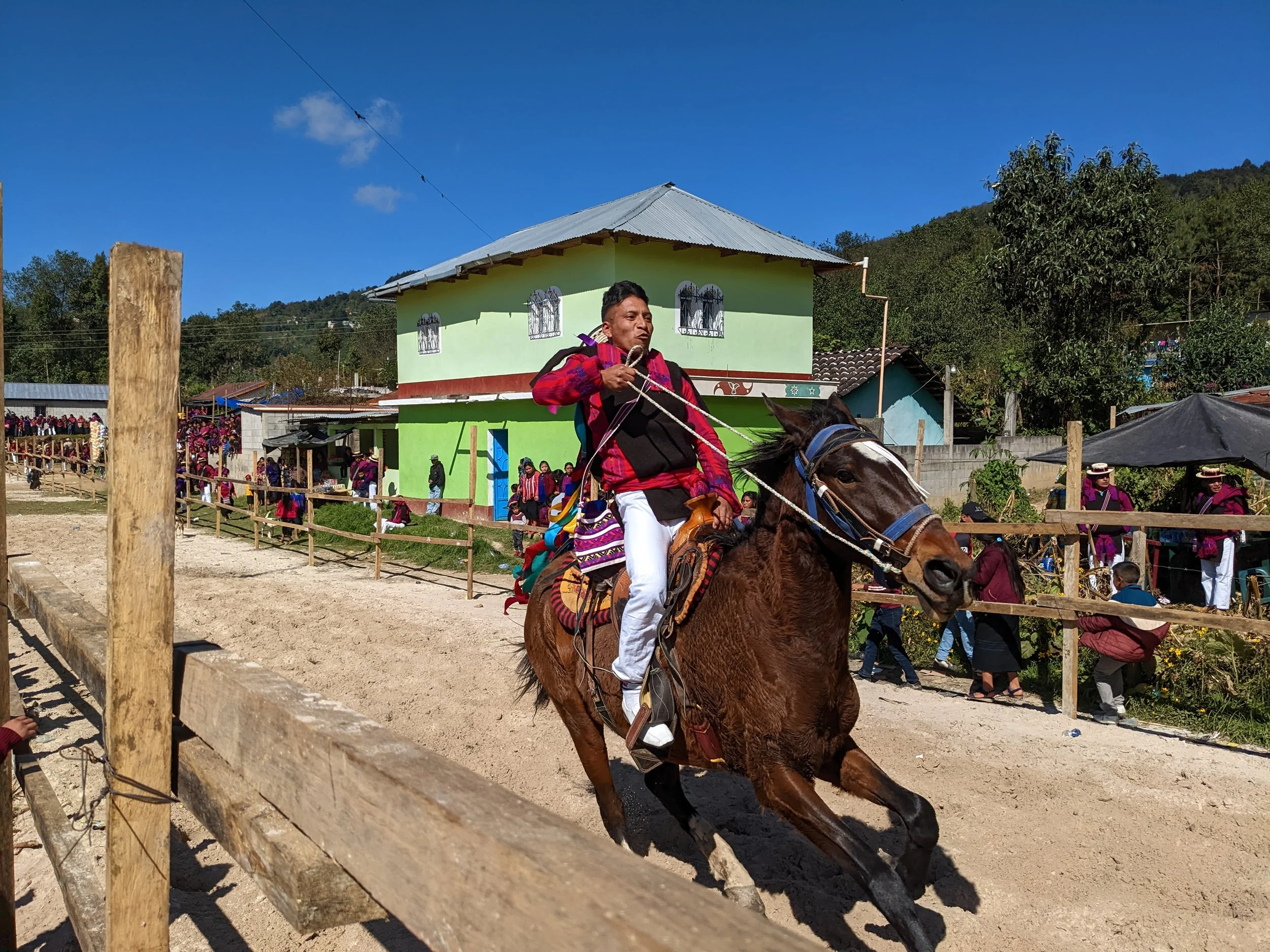 Man wearing traditional clothing riding a horse during a cultural event in a rural area with a crowd of spectators and a green building in the background.