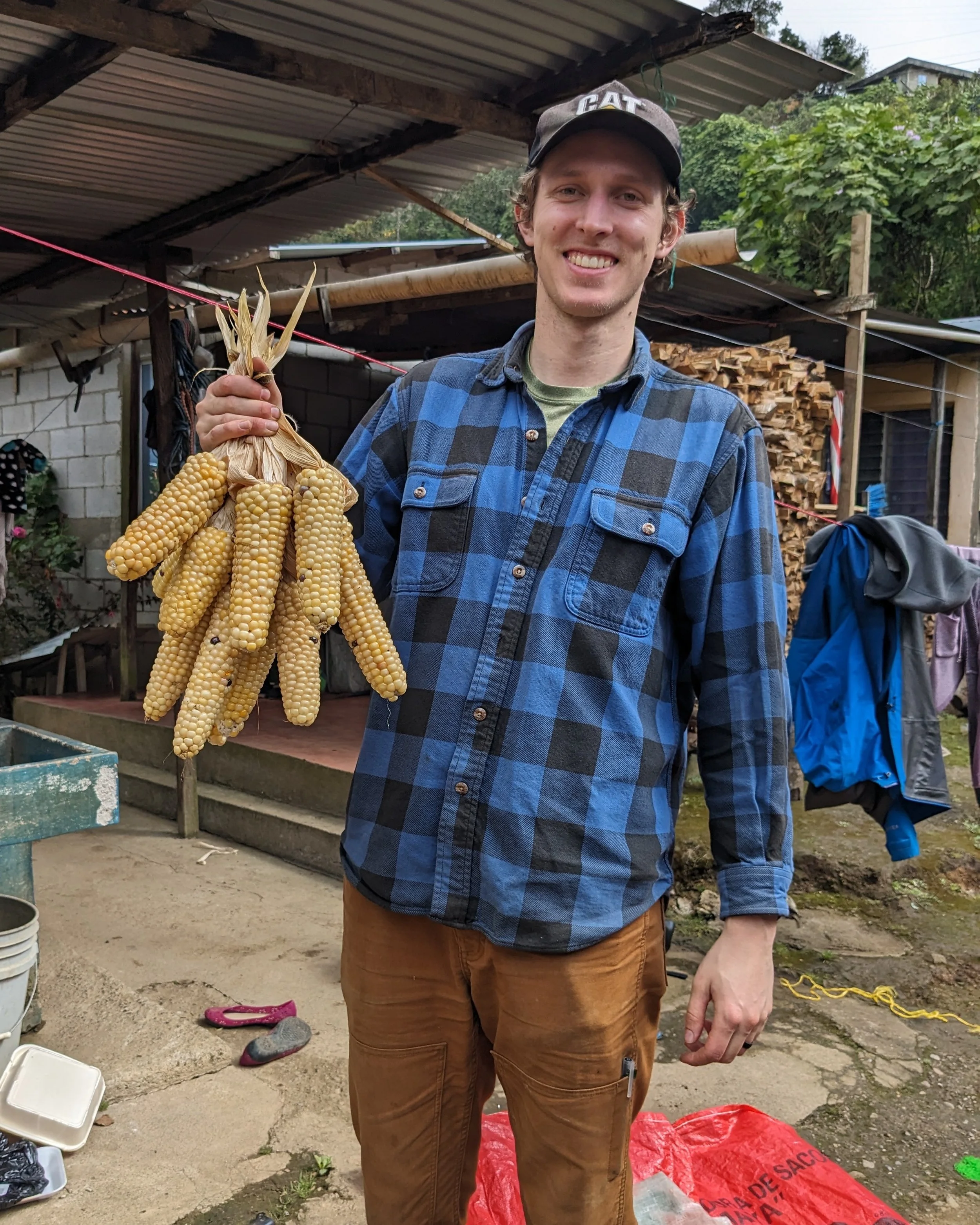 A man wearing a black cap, a blue and black checkered shirt, and brown pants holding a bunch of yellow corn cobs with dried husks in an outdoor setting. There are wood piles, a makeshift shelter, and various objects like shoes, buckets, and a red bag in the background.