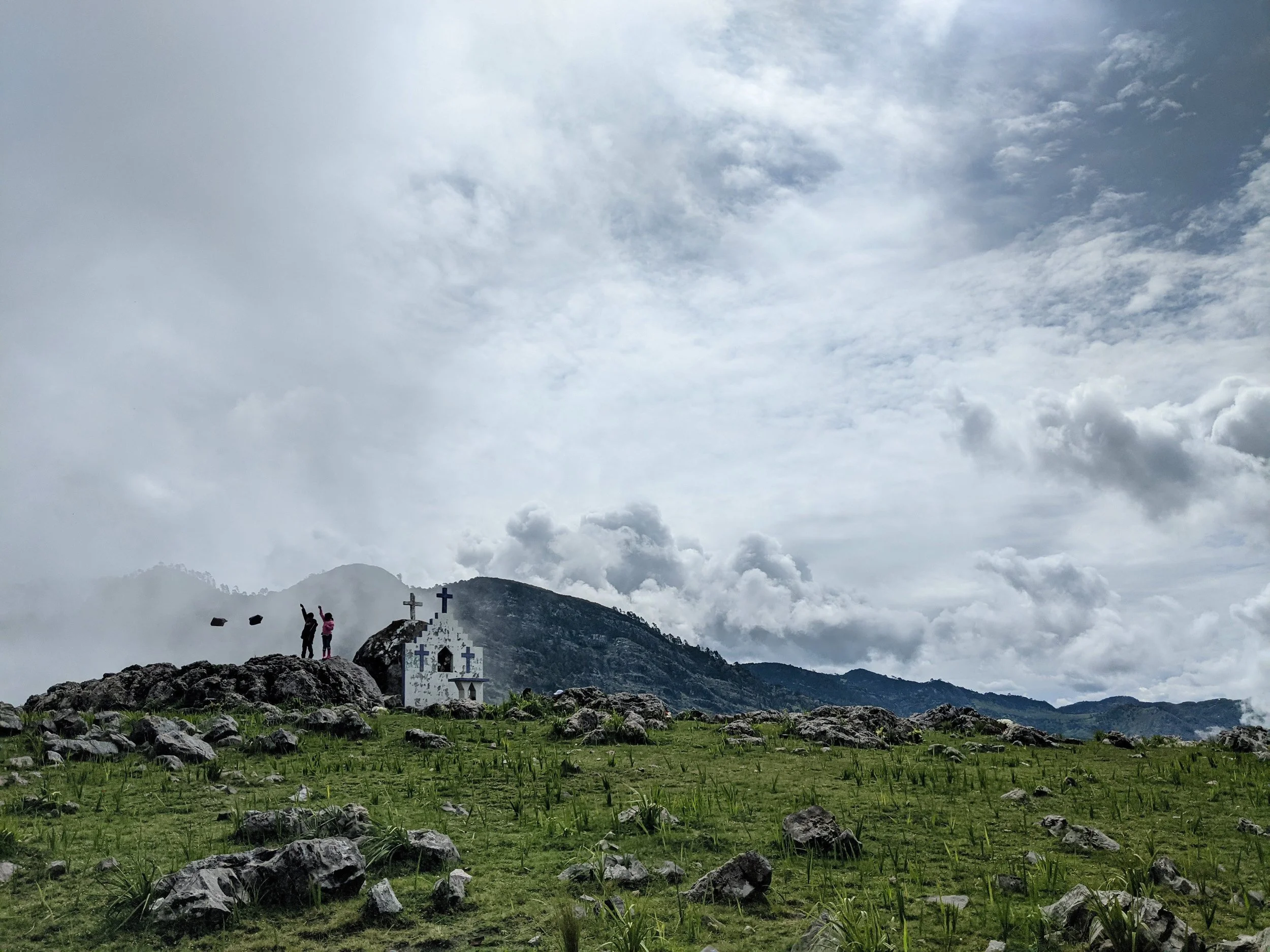 Mountain landscape with rocks and grass in foreground, small white chapel with crosses on its roof, and hills in the background under a cloudy sky.