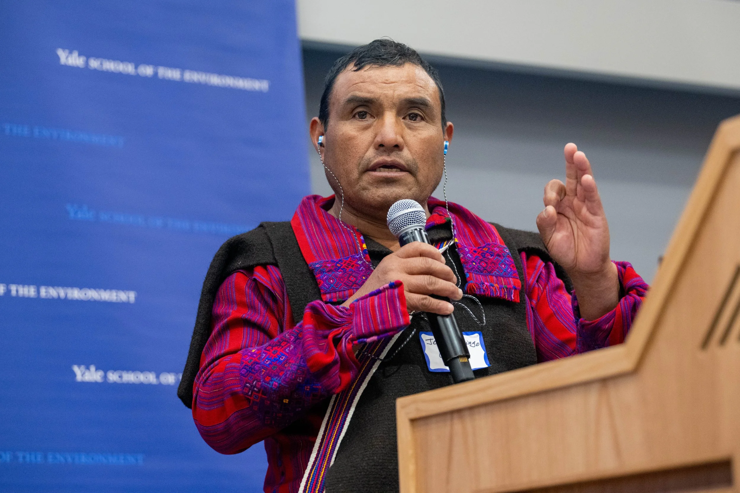 A man speaking into a microphone at a lecture or presentation, wearing traditional Indigenous clothing, with a blue banner in the background.