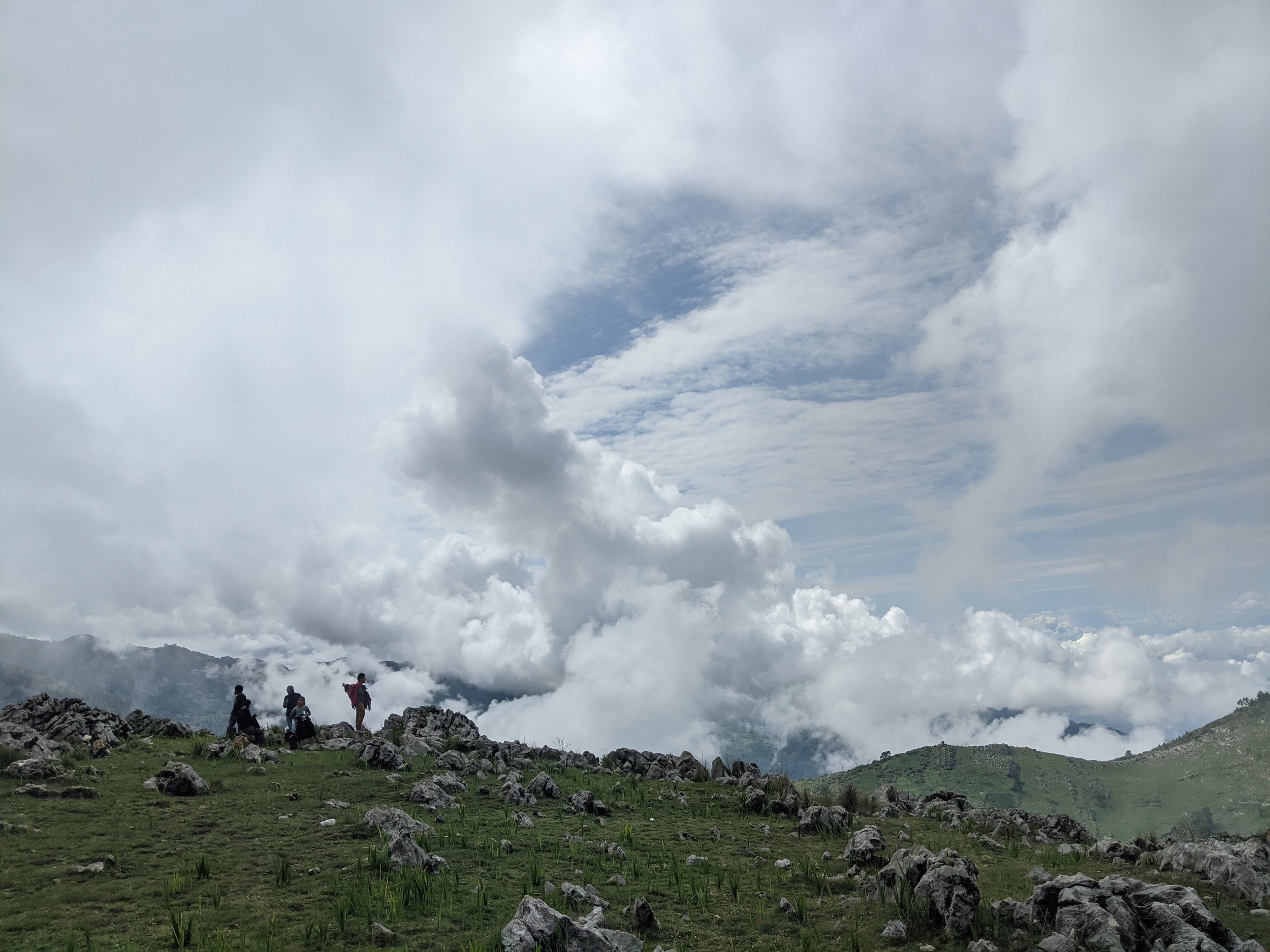 Hikers resting on rocky terrain near a mountain landscape with clouds covering the sky.