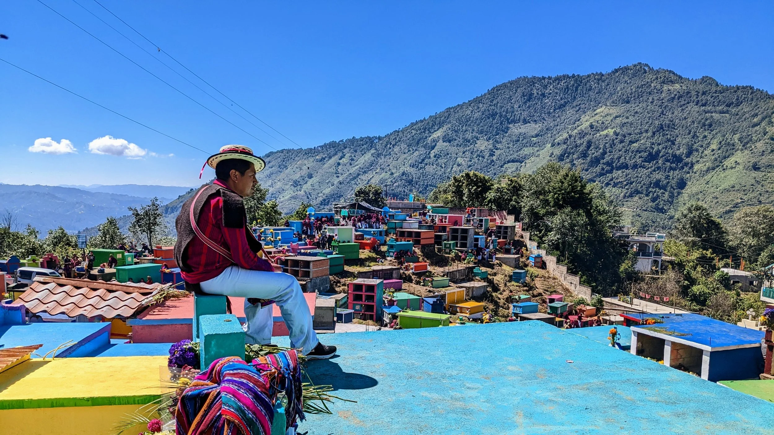 A man in traditional clothing sitting on a colorful rooftop overlooking a hillside cemetery with colorful gravestones, surrounded by green mountains under a clear blue sky.