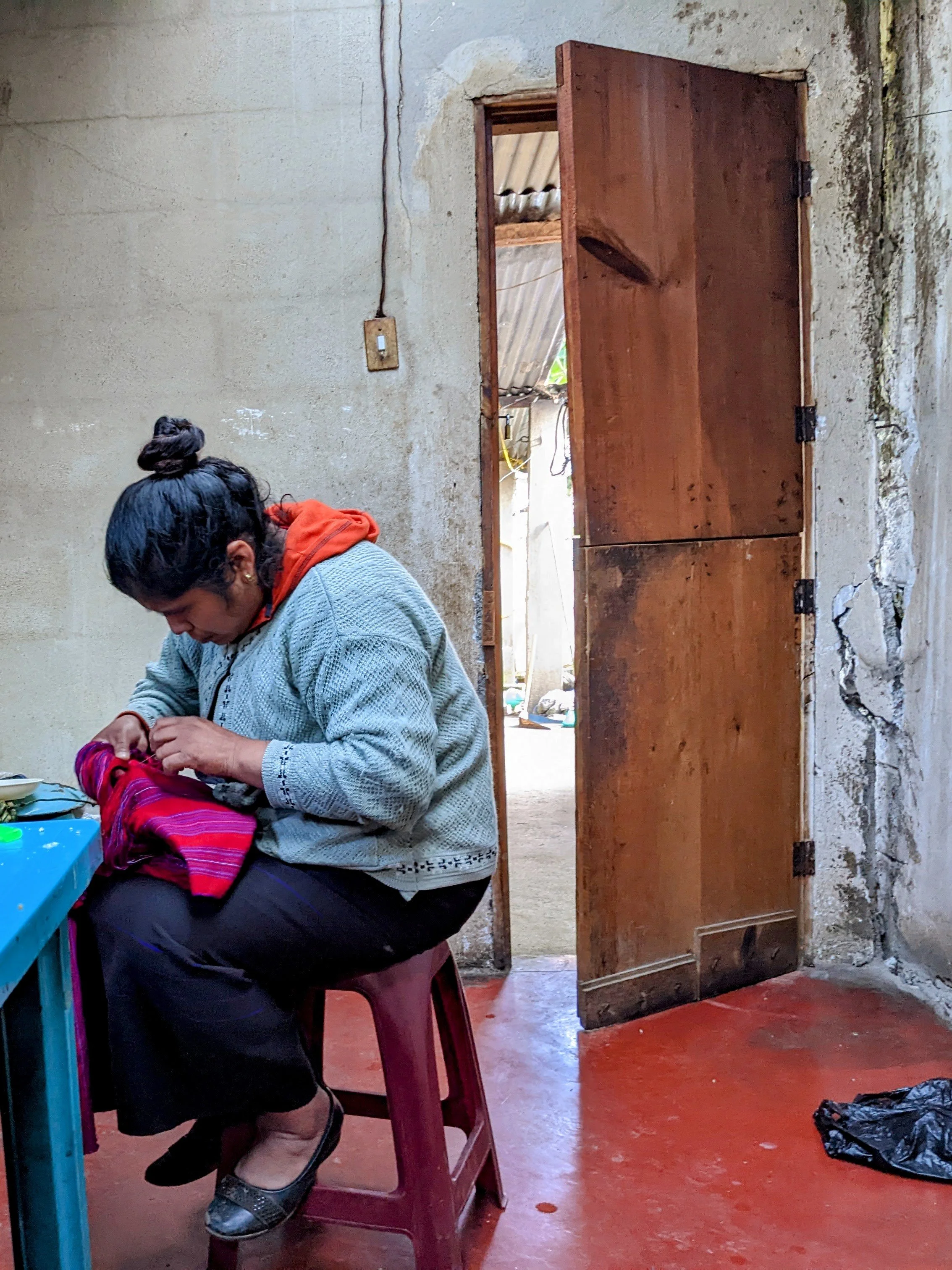 A woman sitting at a table with a pink bag, inside a room with a partially open wooden door, red painted concrete floor, and a wall with an electrical outlet and visible crack.