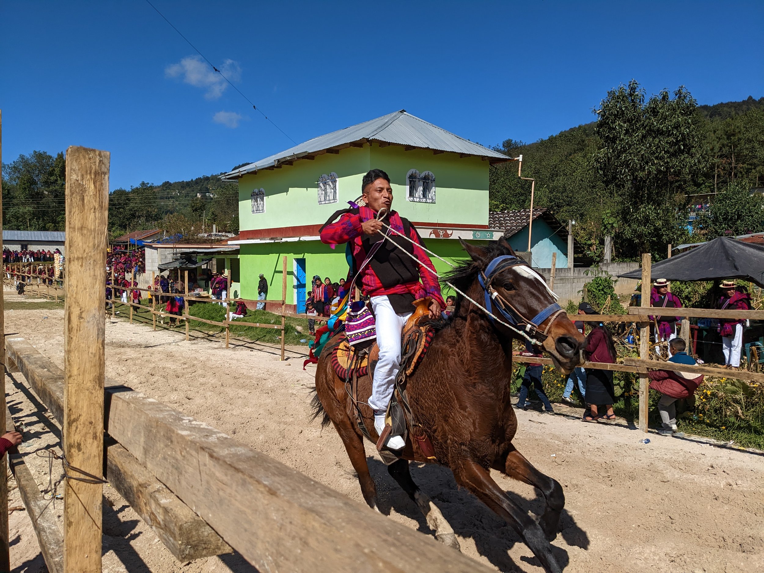 A man riding a horse in a rural setting during a cultural event, with a crowd of people in traditional clothing and a green house in the background.