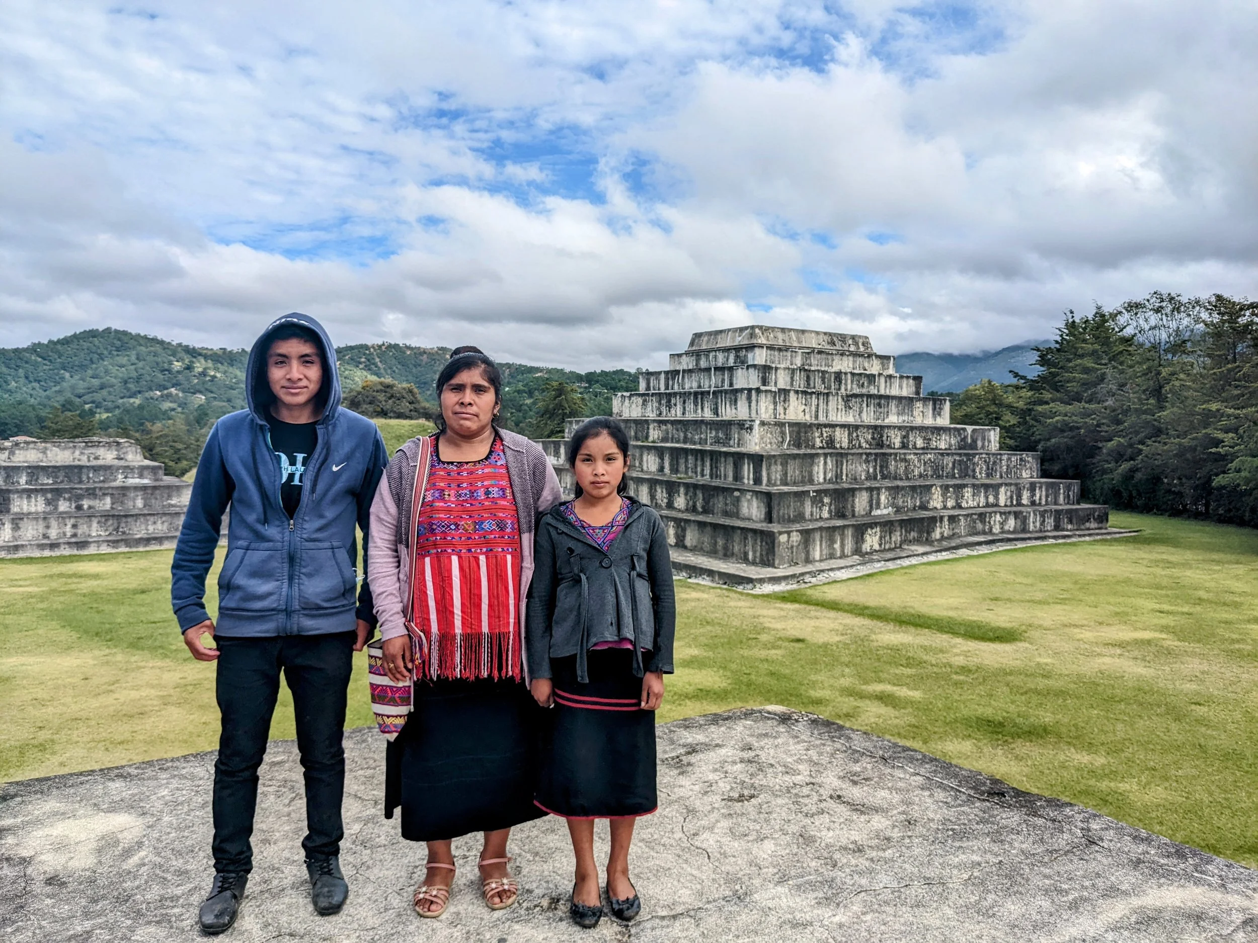 A group of four people standing outdoors in front of ancient Mayan pyramids with mountainous landscape and cloudy sky in the background.