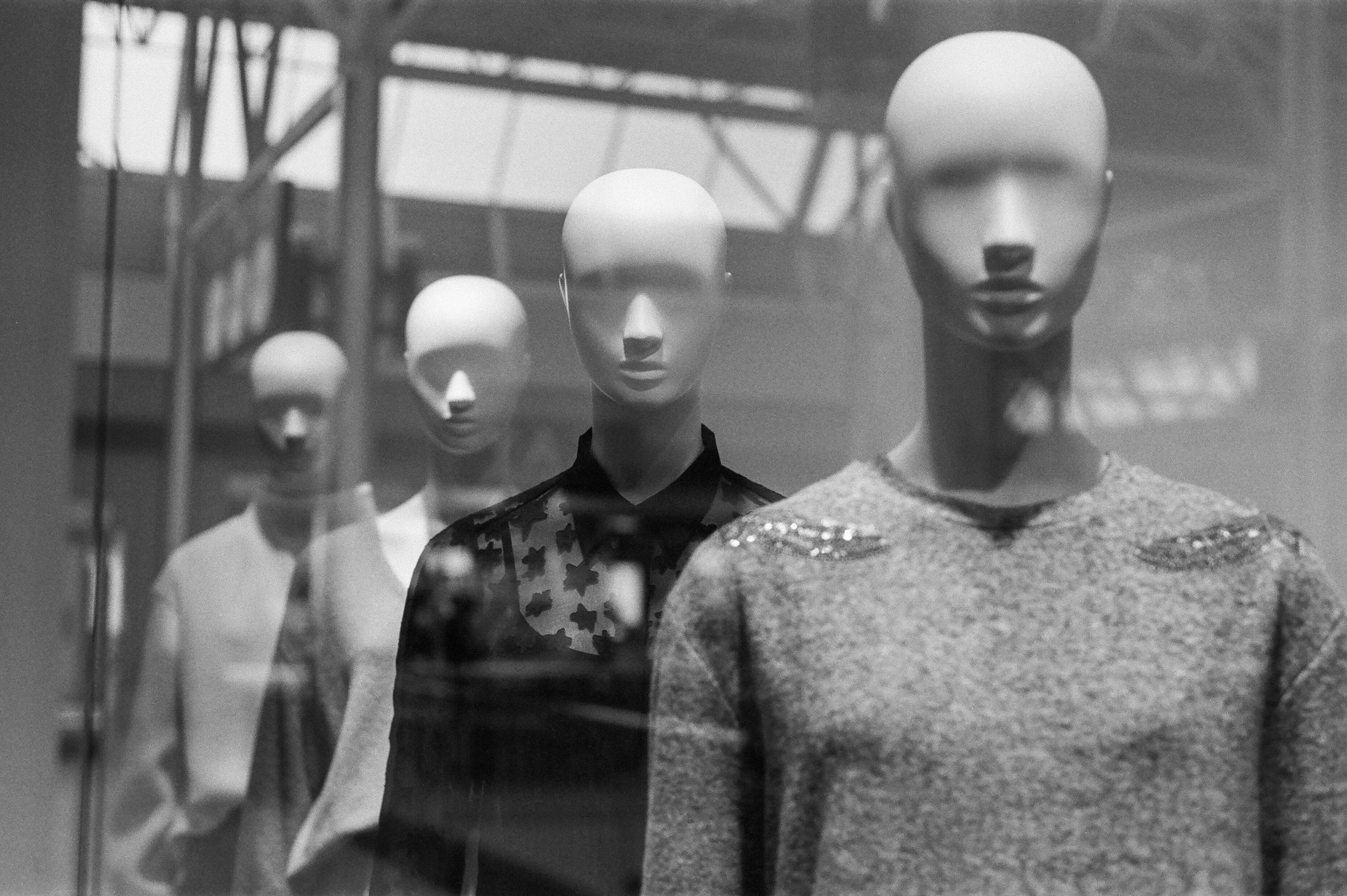 A display window showing mannequins with various outfits, including a sweater with embellishments, a camouflage pattern top, a shirt with a star pattern, and a plain shirt, in what appears to be a retail store.