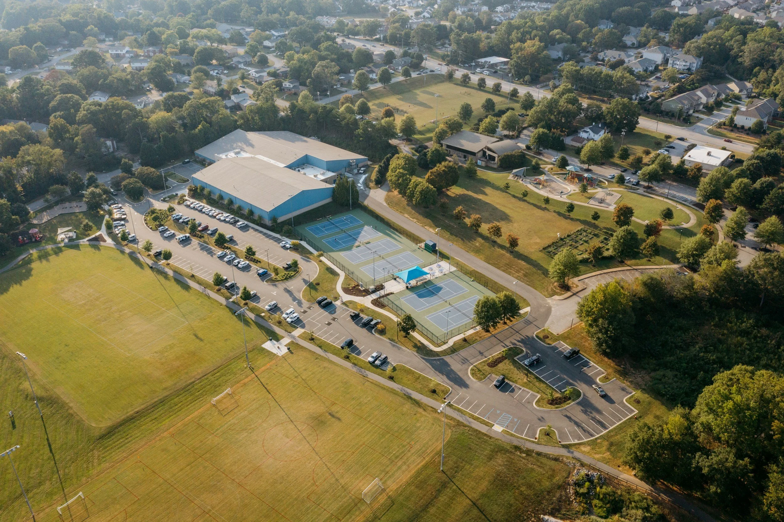 Aerial view of a community park with tennis courts, playground, sports fields, parking lots, and surrounding residential neighborhood with trees.