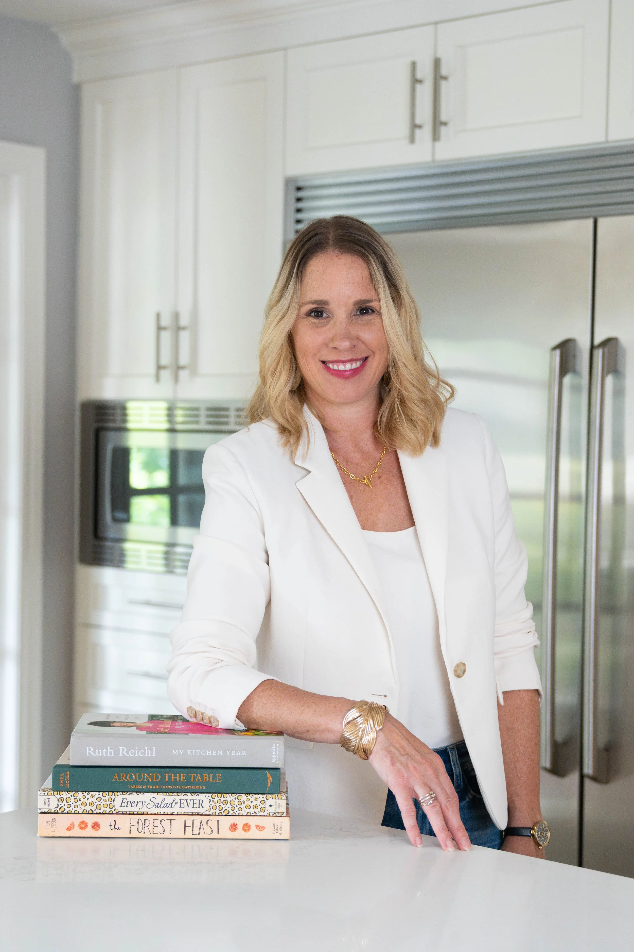 A woman with blonde hair wearing a white blazer and gold jewelry standing in a kitchen. There are stacked cookbooks on the counter in front of her, and a refrigerator is visible in the background.