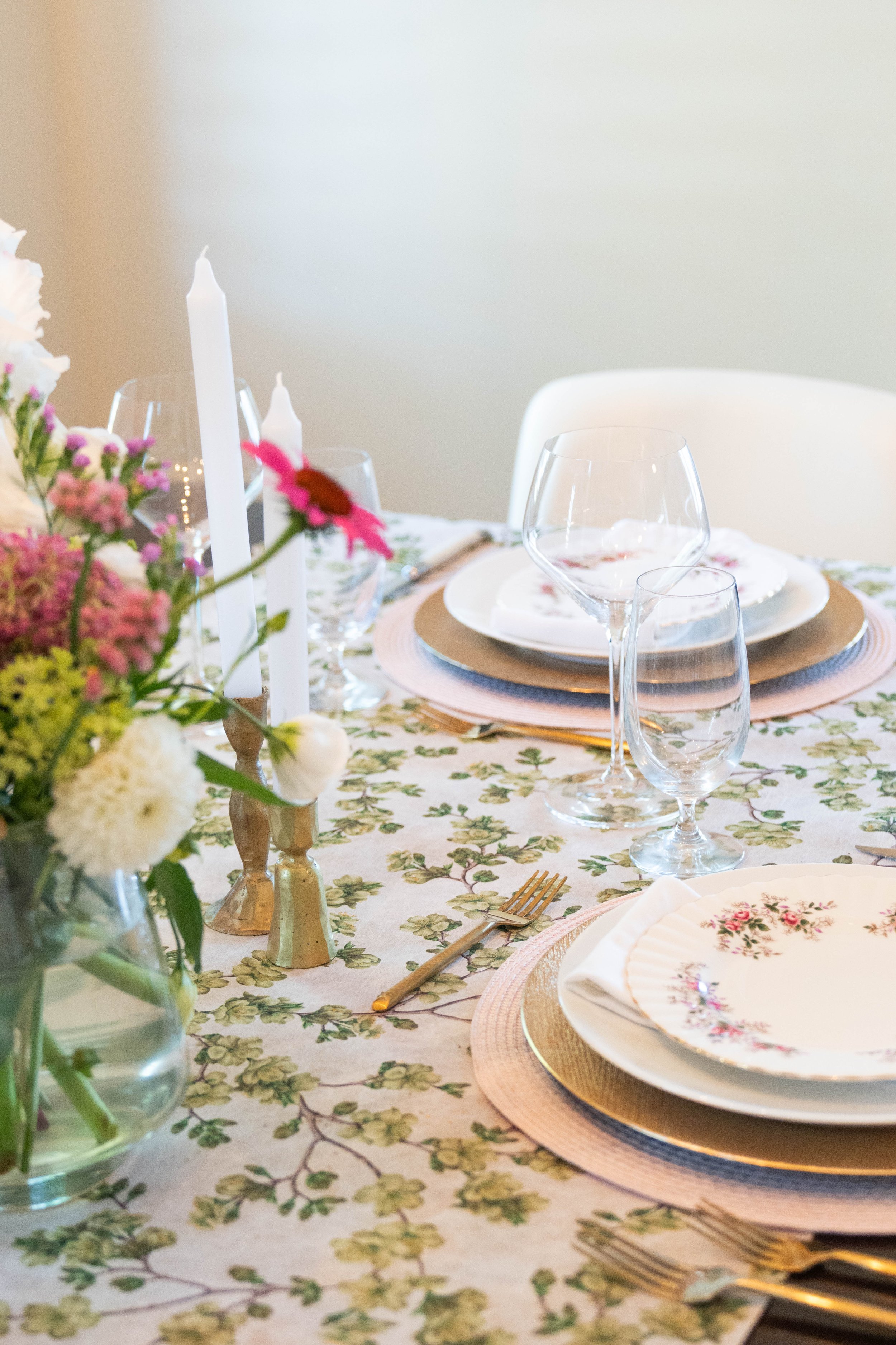 A table setting with a decorative plate featuring pink flowers, gold cutlery, and a floral centerpiece in a gold vase with assorted flowers and greenery.