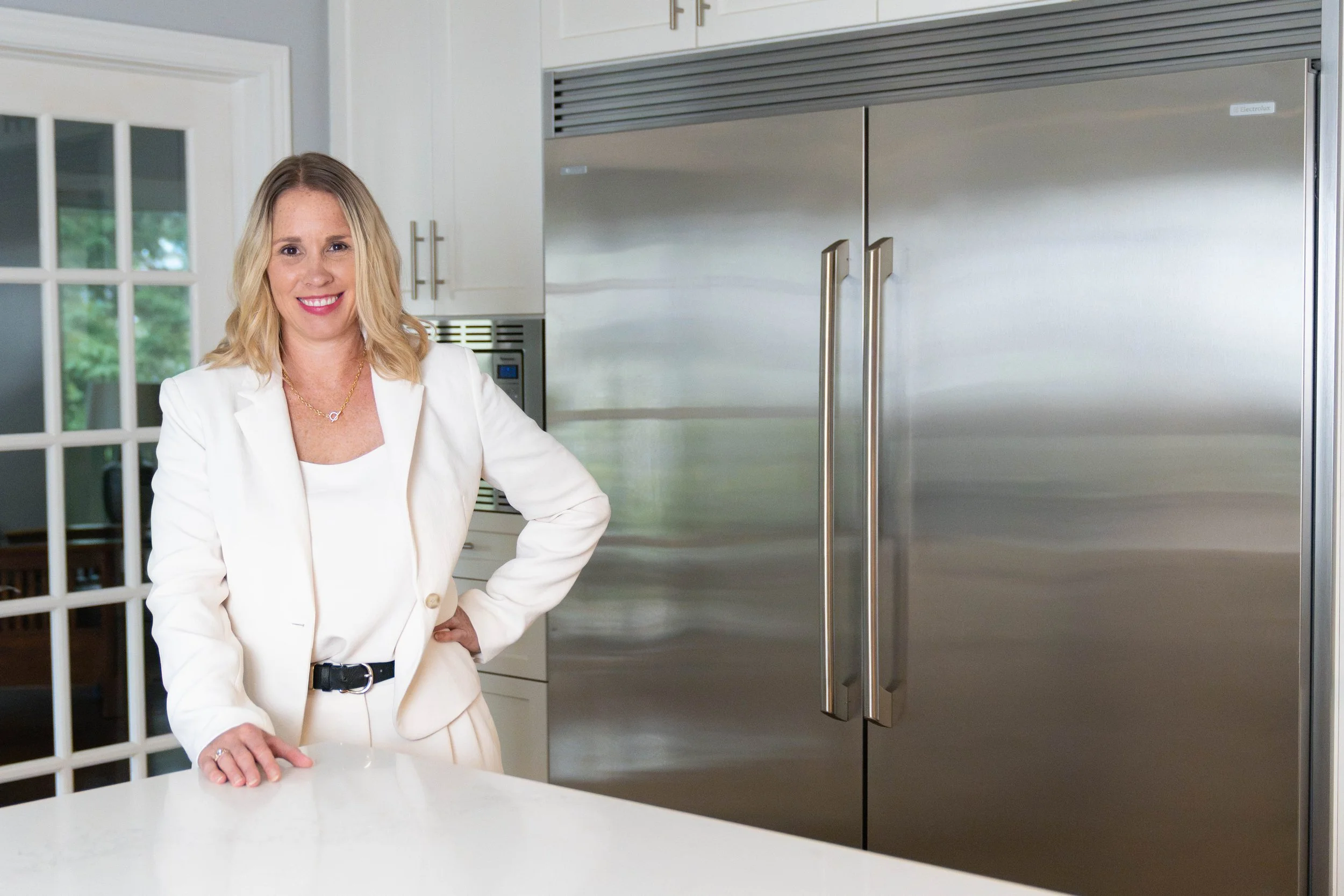 A woman with blonde hair, wearing a white blazer, smiling and standing with her hand on a white table in a modern kitchen with stainless steel refrigerator and white cabinetry.