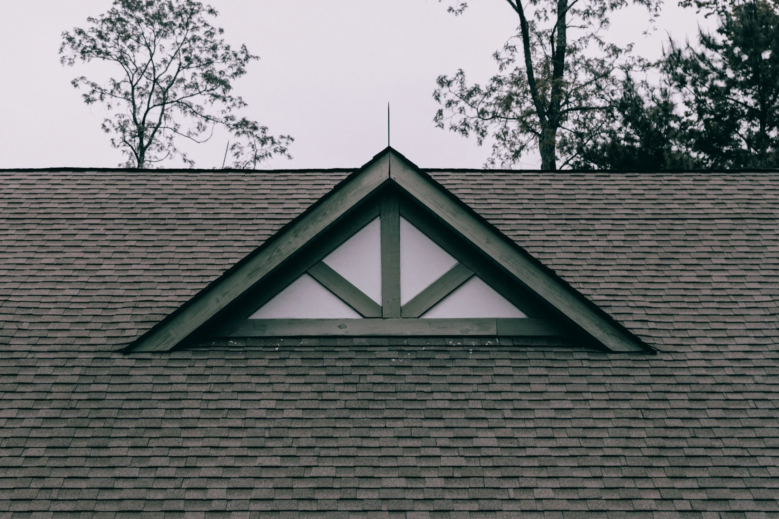 Close-up of a house roof with brown shingles and a triangular gable end, decorated with white and green trim, with trees and an overcast sky in the background.
