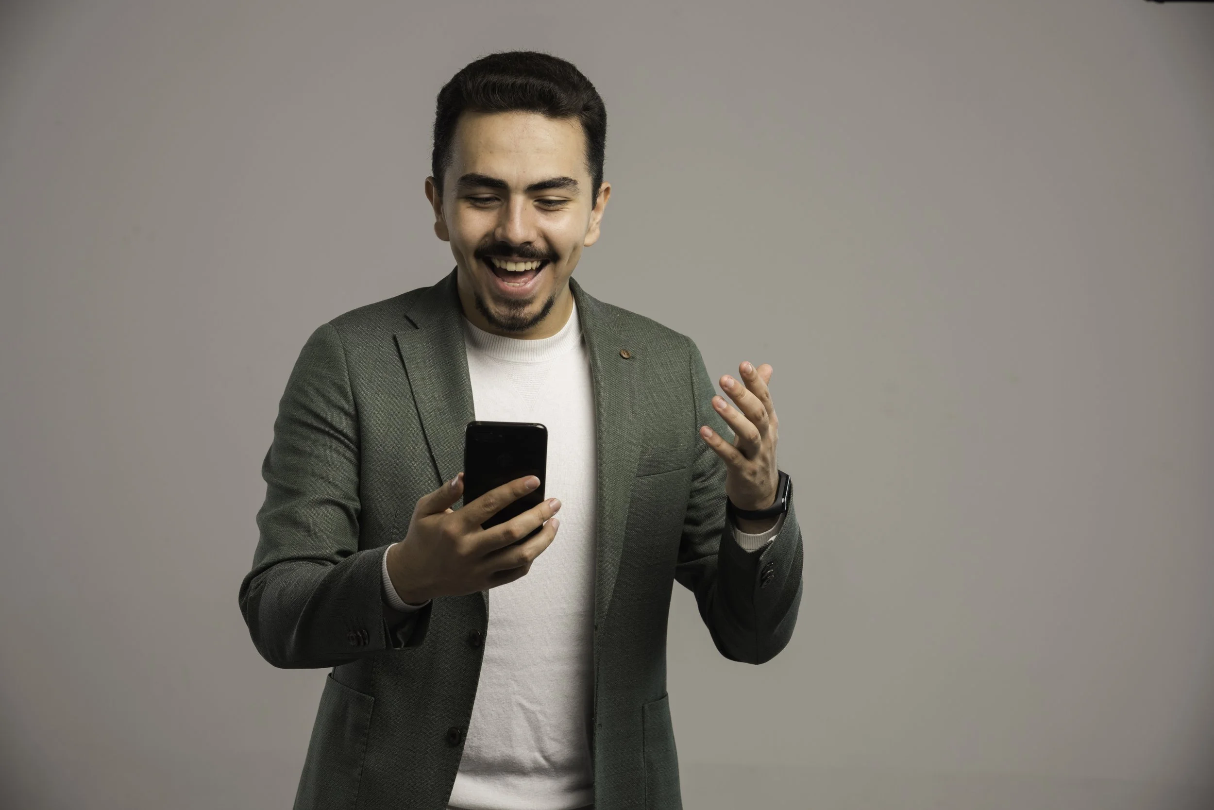 A young man with dark hair and a mustache smiling while looking at his smartphone against a plain gray background.