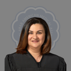 Professional woman with shoulder-length brown hair wearing a black medical gown, standing in front of a gray background with a circular logo that says 'Arab American Medical Association'.