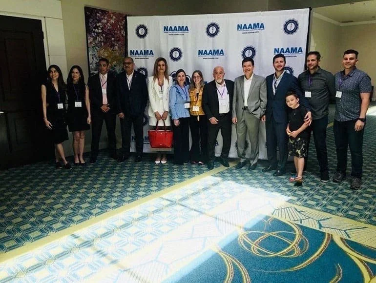 Group of people posing for a photo in front of a NAAMA-branded backdrop at a professional event or conference.