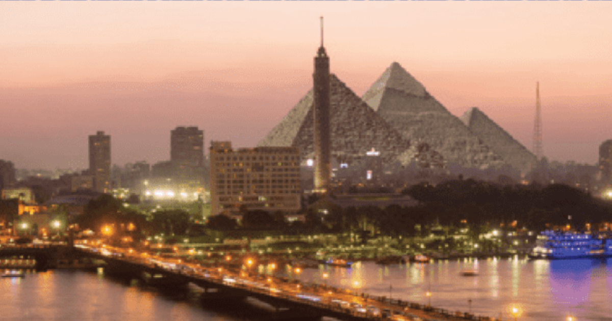 A city skyline at sunset with pyramids, a tall tower, and buildings along a river.