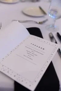 A menu on a white table at a restaurant with cutlery, a glass of water, and a plate in the background.