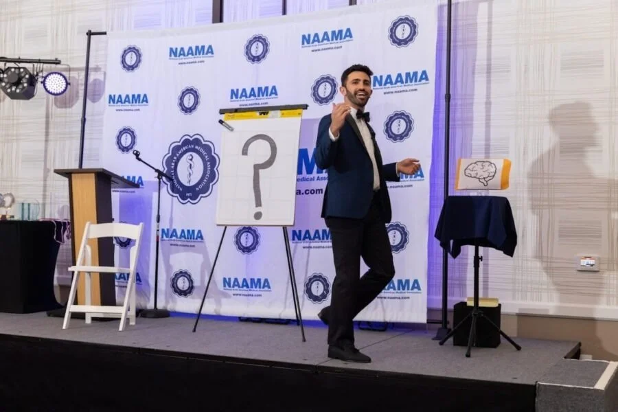Man in formal attire standing on stage with NAAMA logo background, gesturing with one hand, near a large whiteboard with a question mark, and a small stand with a diagram of a brain.
