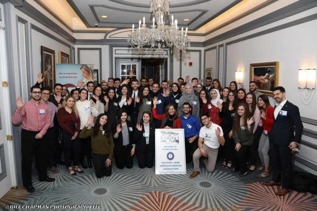 Group of diverse young professionals and students posing indoors at a conference or event, with a sign for the National Arabs American Medical Association in front.