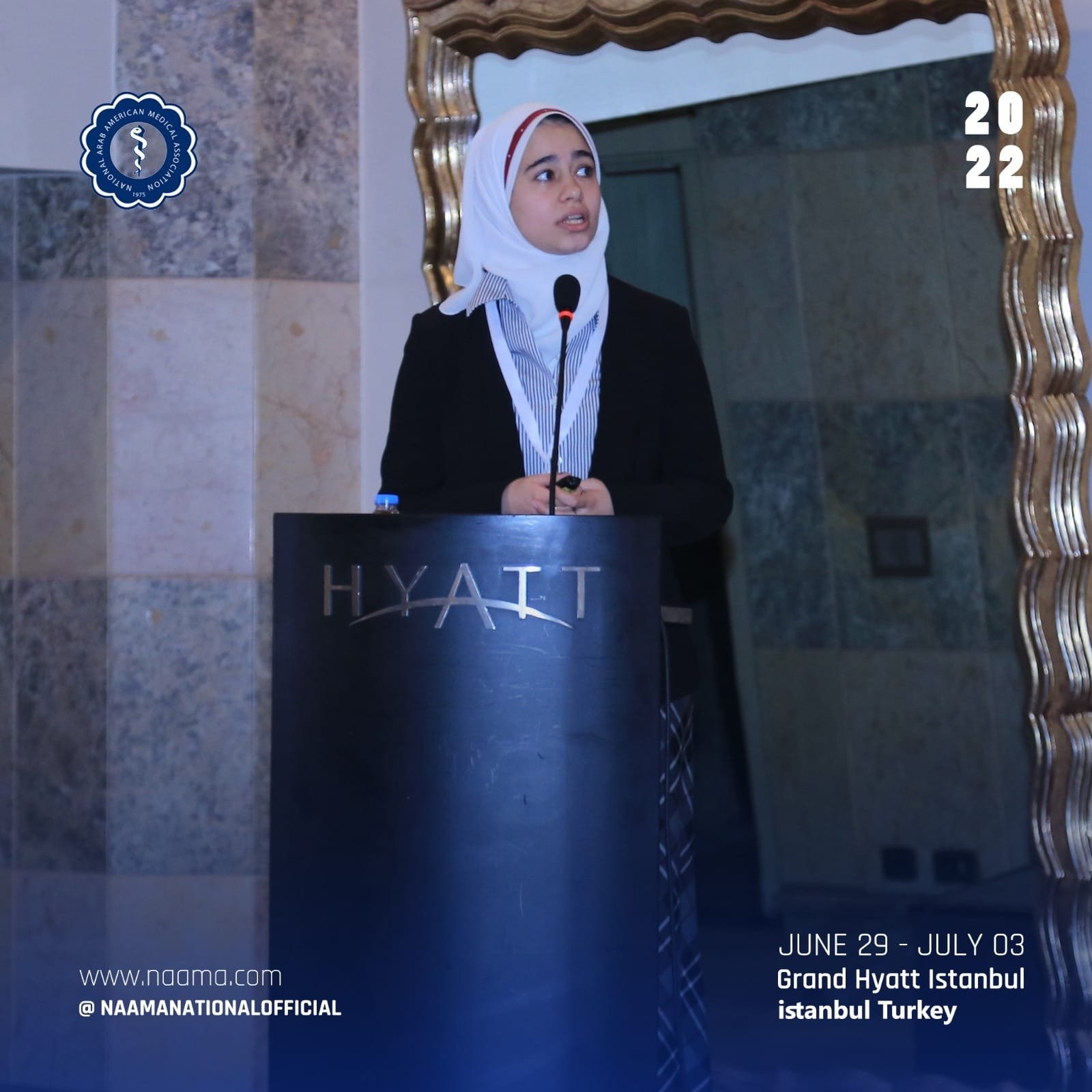 A young woman wearing a white hijab and black blazer standing at a lectern with a microphone at the Grand Hyatt Istanbul. A mirror with a gold frame is behind her. The event is from June 29 to July 3, 2022, organized by the Arab American Medical Asso