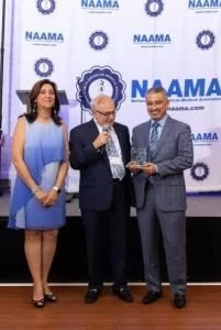 Three people standing in front of a backdrop with NAAMA logos. The woman on the left is wearing a blue dress, the man in the middle is wearing a dark suit and glasses, and the man on the right is in a gray suit holding an award or plaque.