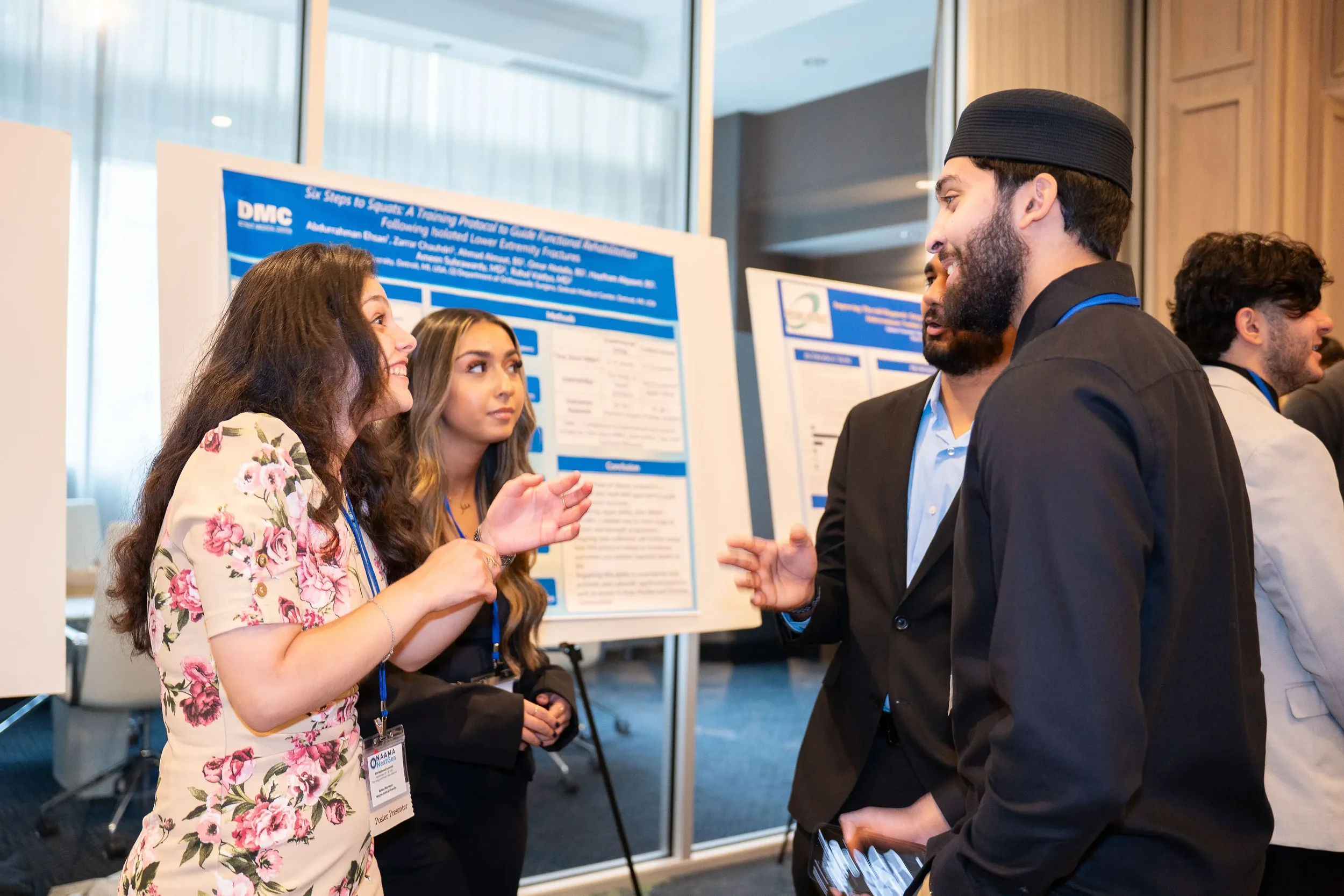 A group of people at a conference or professional event, engaging in conversation. Two women and one man are in the foreground, with a poster presentation in the background displaying scientific or academic information.