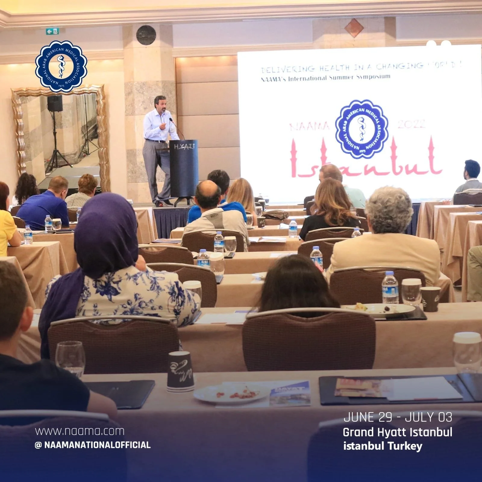 A man in a white shirt and gray pants is standing at a podium with a microphone, giving a presentation at a conference room. Behind him, a large screen displays a slide with the NAAMA logo, the event slogan, and the dates June 29 - July 03 in Istanbu