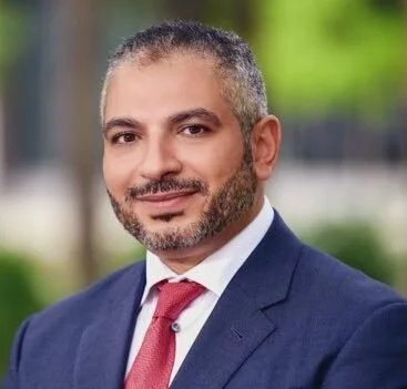 Headshot of a man with short gray hair and a beard, wearing a navy suit, white shirt, and red tie, smiling outdoors.