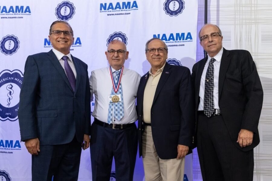 Four men in suits standing together at an event with NAAMA logos in the background, one man wearing a medal around his neck.