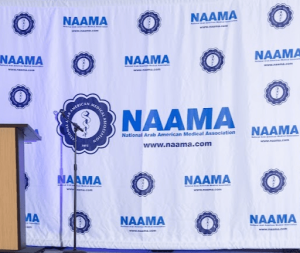 Blue and white NAAMA backdrop with logo and repeating pattern behind a podium.