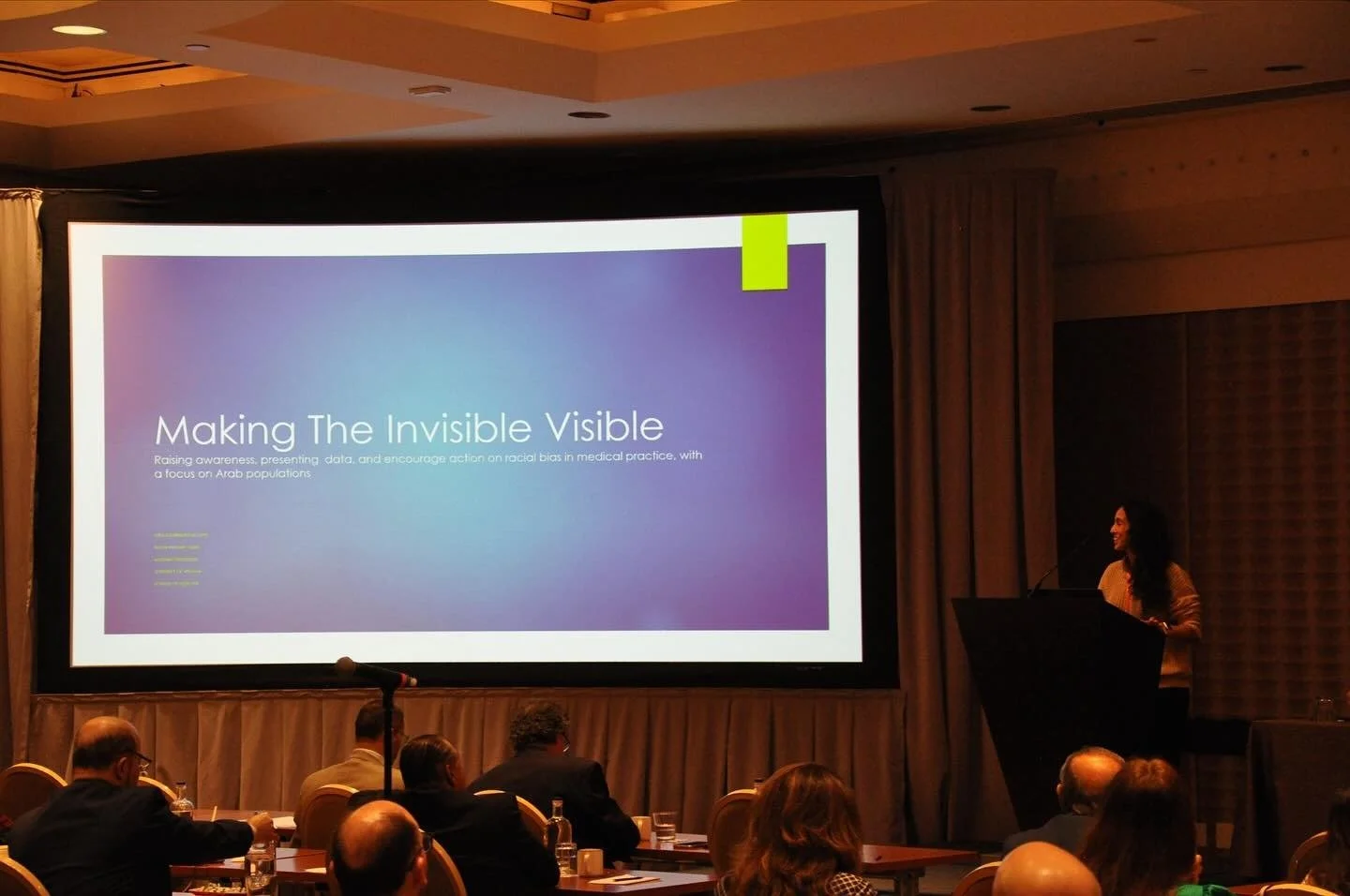 A woman giving a presentation at a conference with a large screen showing the title 'Making The Invisible Visible' and an audience seated in front.
