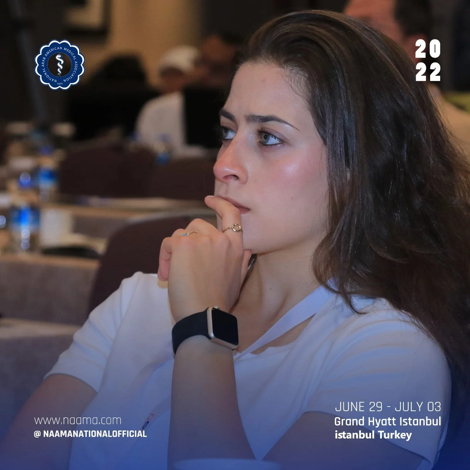 Close-up photo of a woman with long brown hair, blue eyes, and light skin, wearing a white top, silver rings, and a smartwatch. She is seated at an indoor event, with a blurred background of other people and water bottles. The overlay text indicates 