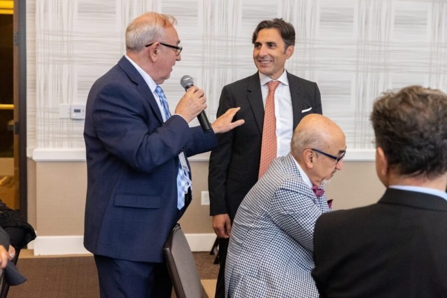 Four men in business attire engaging in conversation at a formal event. One man is speaking into a microphone, while the others listen and smile. The setting appears to be a conference room or banquet hall.