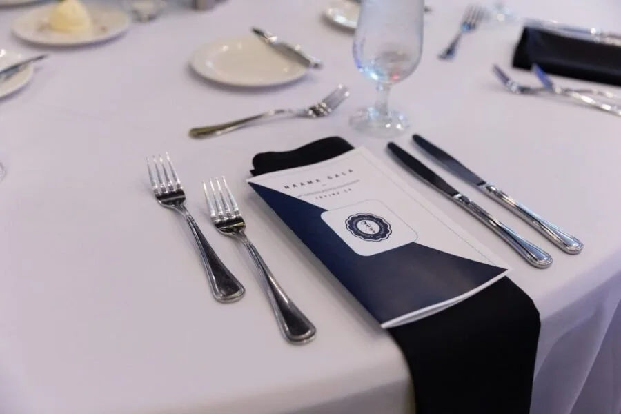 Table setting with silverware, a menu, a black napkin, and a water glass on a white tablecloth.