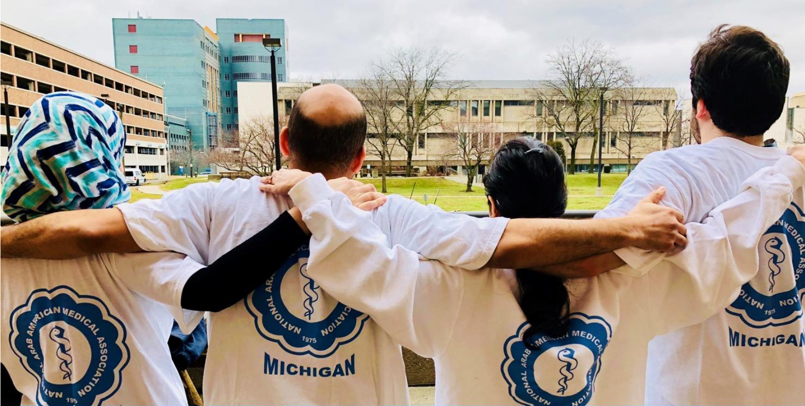 Group of diverse medical professionals standing outdoors with arms around each other, wearing white T-shirts with a logo of the Arab-American Medical Association of Michigan.