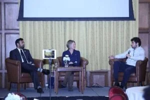 Three people seated on stage during a panel discussion, with a large screen above them and a yellow curtain backdrop.
