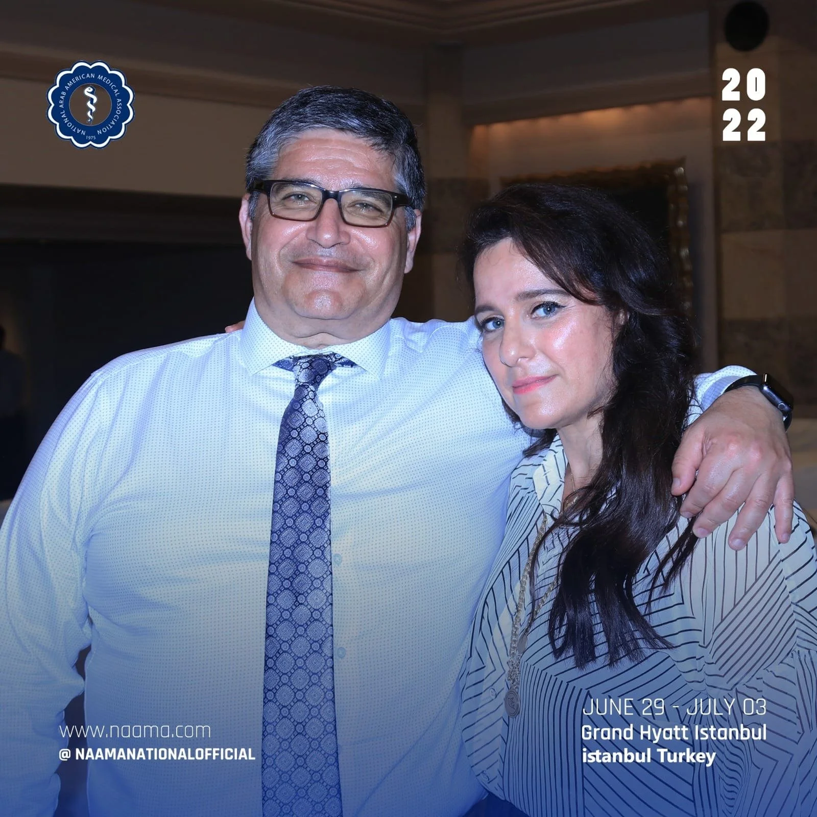 Two smiling people, a man with glasses and a woman with dark hair, are posing together at an event. The man is wearing a white shirt and patterned tie, and the woman is dressed in a striped shirt. They are in an indoor setting with a wooden wall back