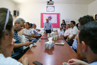 Medical staff gathered in a meeting room around a large table, with a woman speaking at the center and a pink and white medical emblem on the wall behind her.