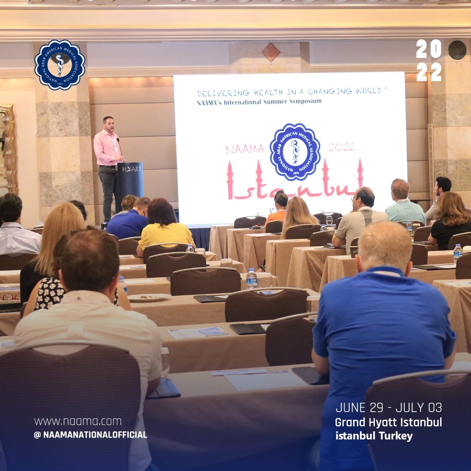 A conference room filled with people sitting at tables, listening to a speaker presenting on a large screen. The slide on the screen displays the NAAMA logo, the event title 'Delivering Health in a Changing World!', and mentions the dates June 29 - J