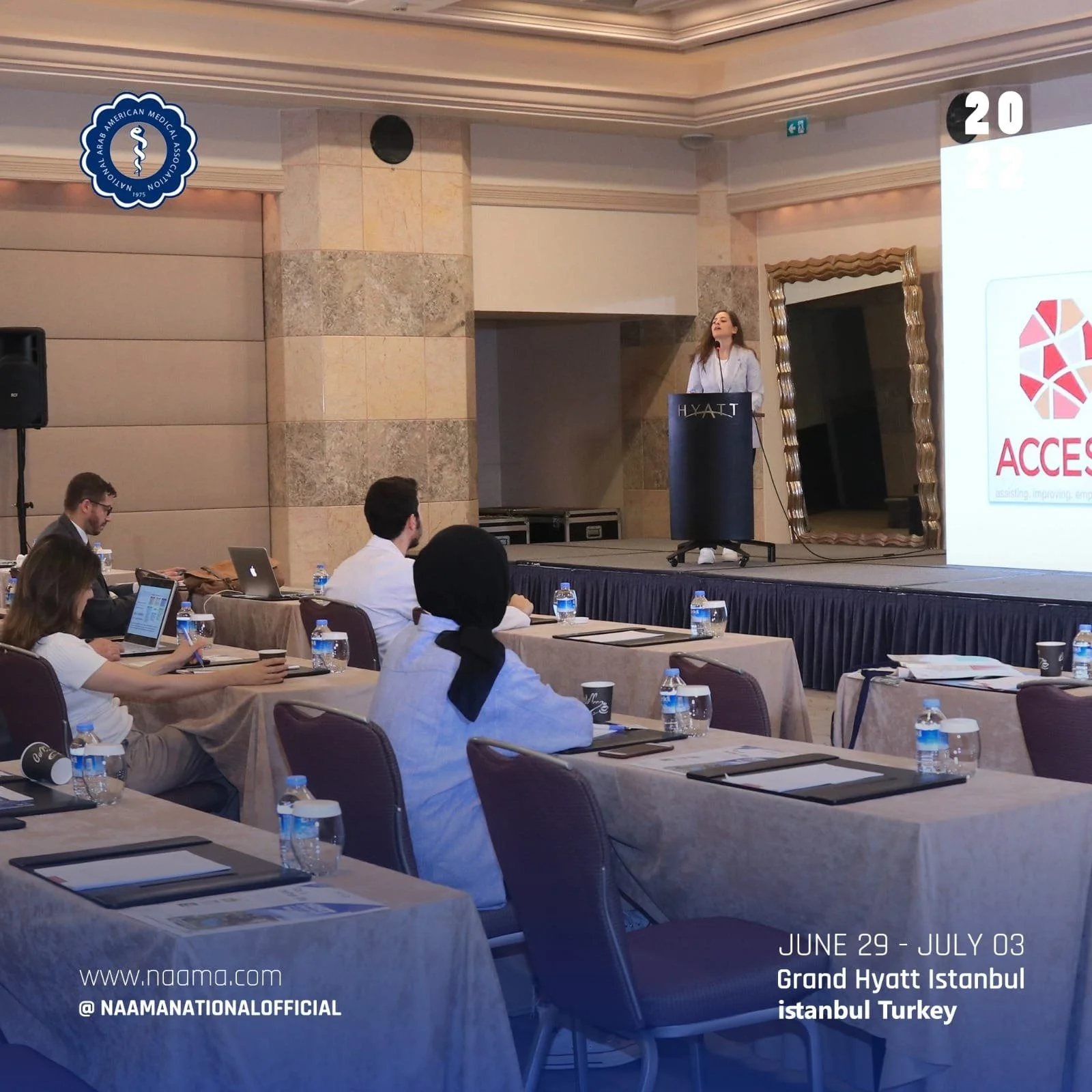 A woman speaking at a conference podium in a large, formal hotel conference room, with attendees seated at tables listening. The room has marble walls and a large screen with the event's logo and details, including dates and location in Istanbul, Tur