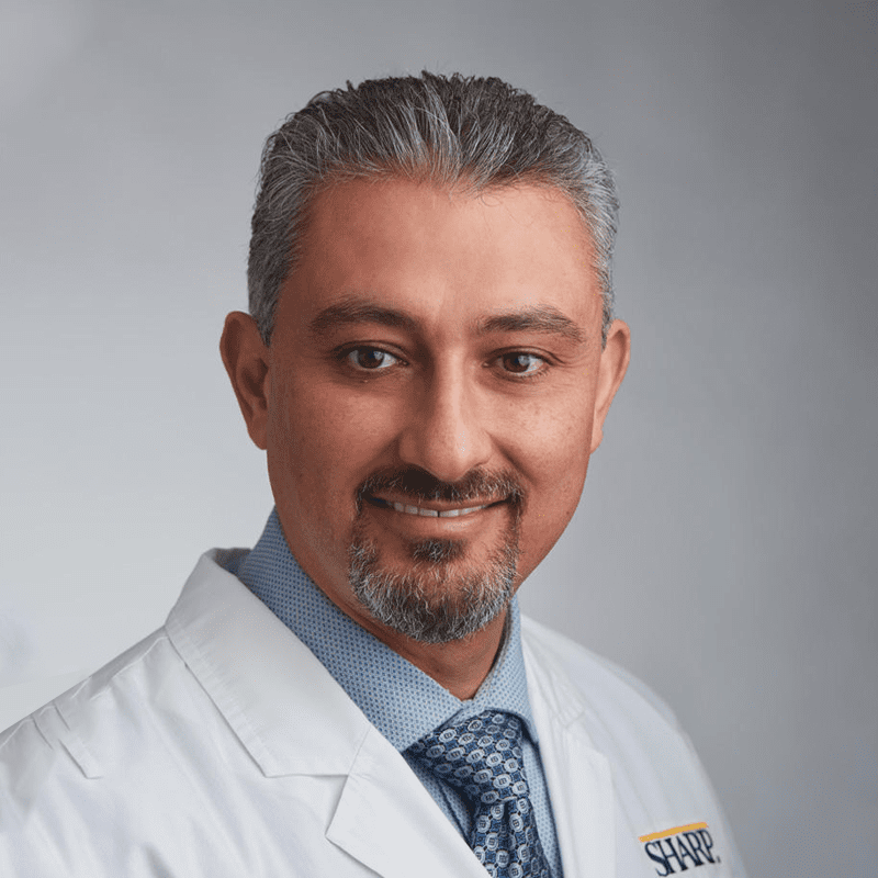 A male doctor wearing a white coat, light blue shirt, and patterned tie, smiling against a plain gray background.