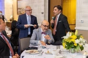 A man in a suit with a microphone speaking to a seated man with glasses and a bowtie at a dining table with flowers.