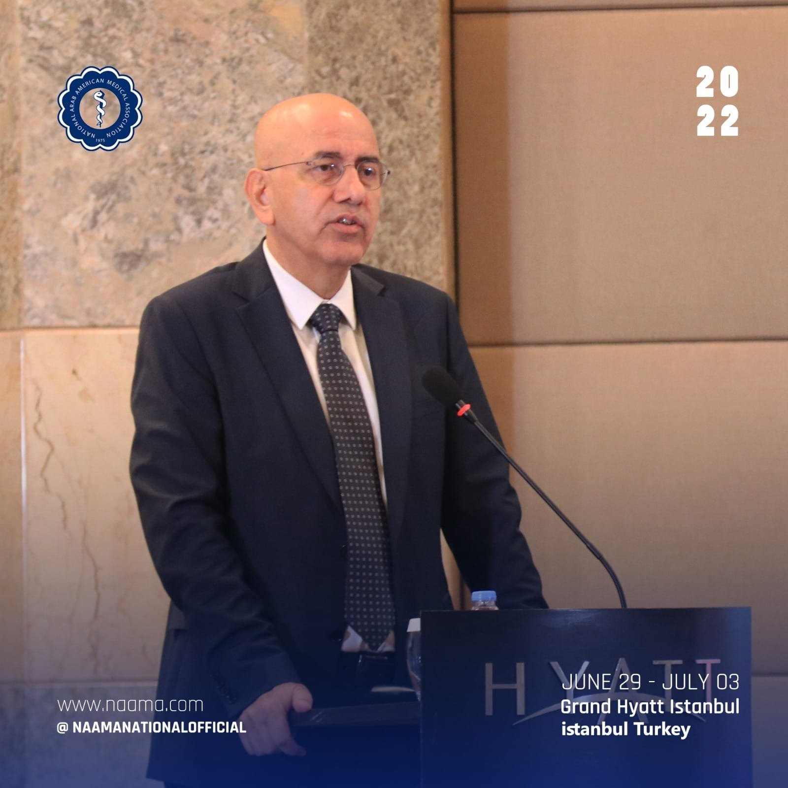 A man in a suit and tie is standing at a podium giving a speech at the NAAM conference at the Grand Hyatt Istanbul in Istanbul, Turkey, from June 29 to July 03, 2022. There is a logo on the wall behind him and text indicating the conference details.