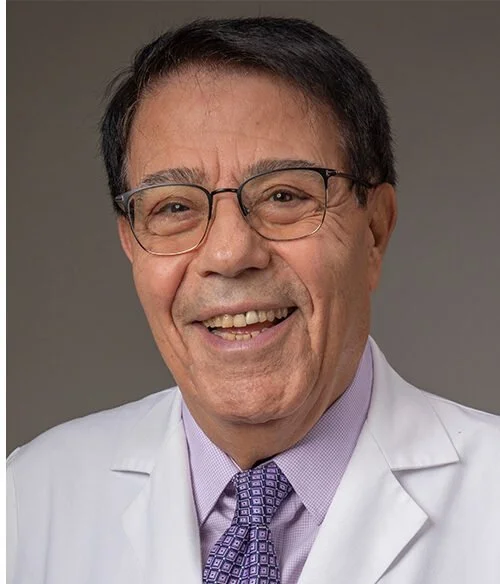 A smiling middle-aged man wearing glasses, a white lab coat, a light purple shirt, and a patterned purple tie, against a plain gray background.